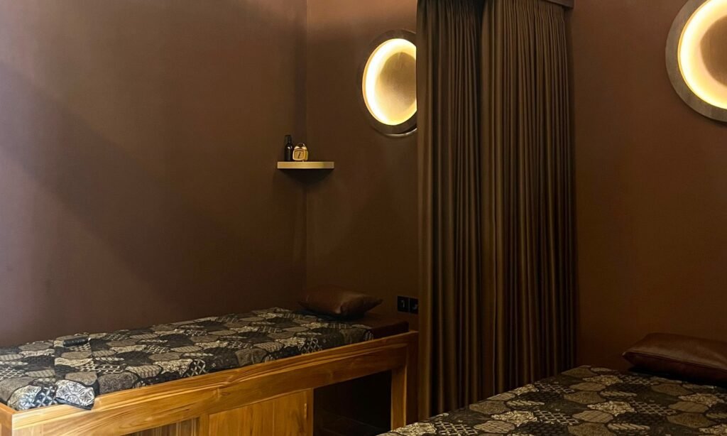 Teak massage bed with batik cover beside a round, warmly lit wall lamp